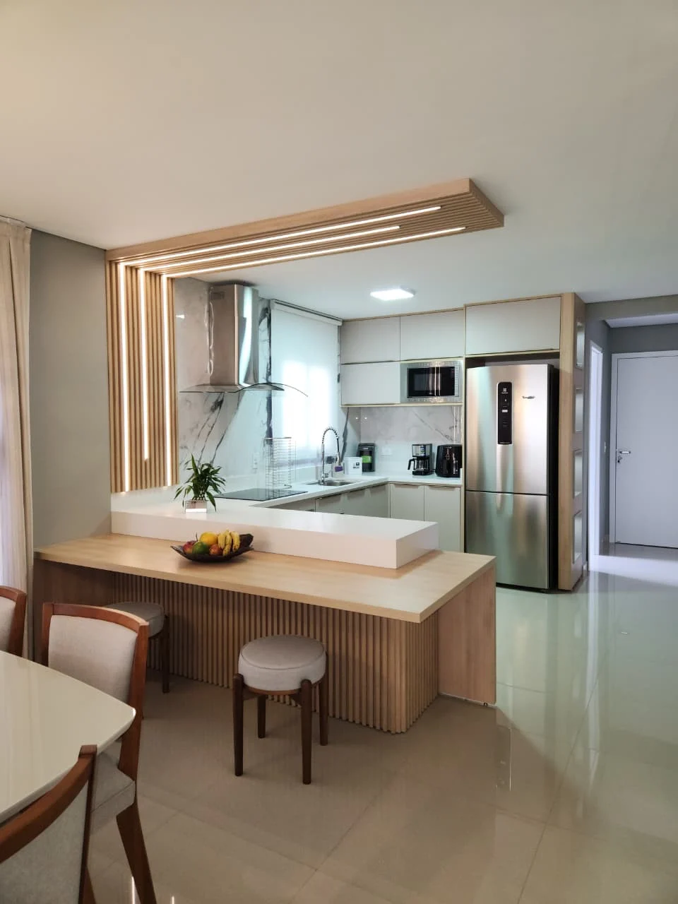 Cozinha sob medida em U com península de frente ripada em madeira clara, bancada em quartzo branco e sanca ripada com iluminação linear embutida