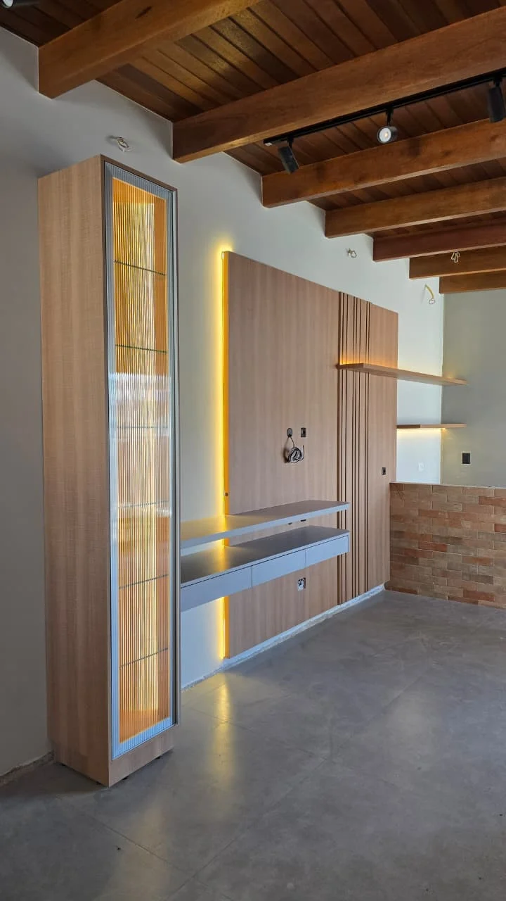 Área gourmet com painel em lâmina de madeira, torre alta com porta de vidro canelado iluminado e bancada suspensa, sob teto de vigas aparentes
