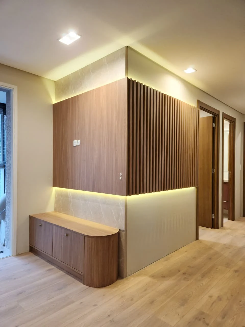 Painel divisor de hall em nogueira, com uma face lisa e outra ripada, iluminação embutida em cima e embaixo e banco baixo com canto arredondado